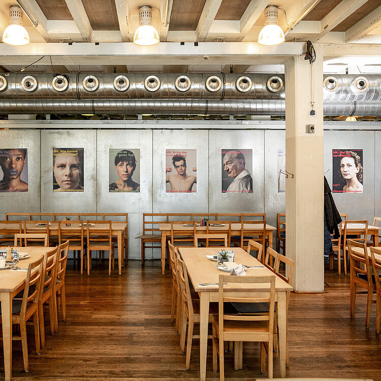 Innenaufnahme eines modernen Restaurants mit Holztischen und -stühlen. An der Wand hängen stilisierte Porträtfotos von Menschen. Die Beleuchtung ist hell, und die Einrichtung wirkt schlicht und einladend.