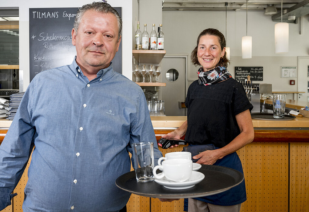 Zwei Mitarbeitende in einem Café: ein Mann hält ein Tablett mit Tassen und Gläsern, eine Frau lächelt im Hintergrund hinter der Theke. Beide wirken freundlich und servicebereit.