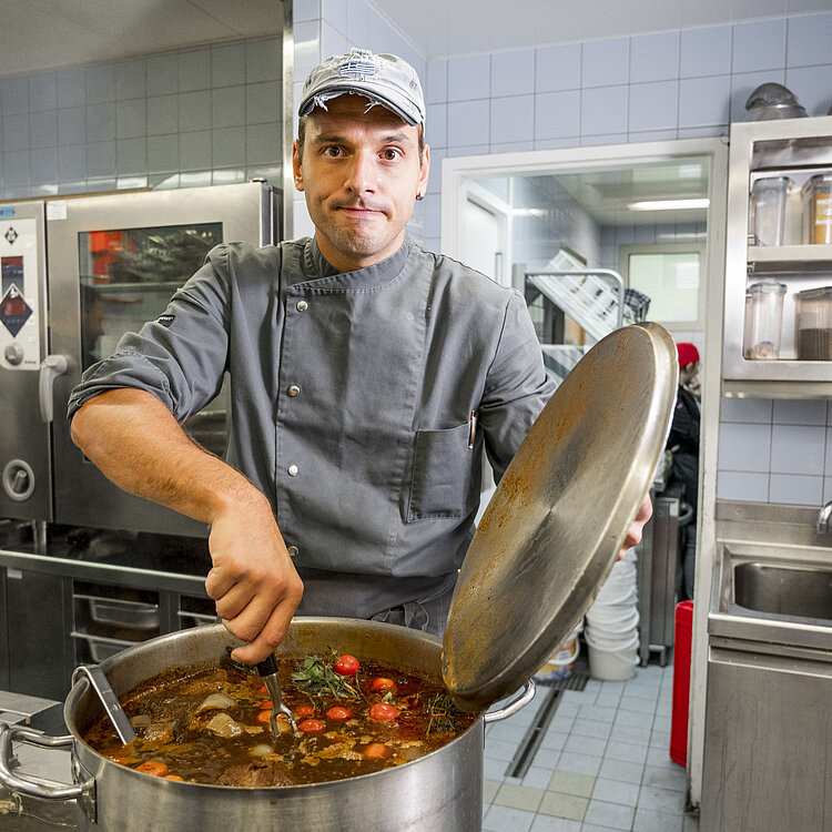 Ein Koch hebt den Deckel eines großen Topfes in einer professionellen Küche und rührt darin eine Suppe mit Gemüse und Fleisch.