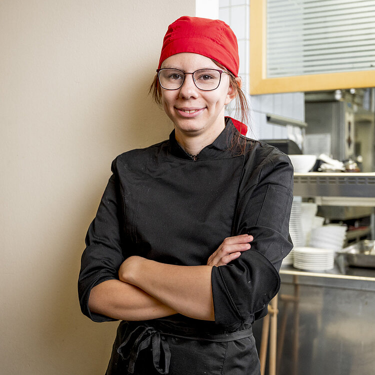Eine Köchin mit rotem Kopftuch und schwarzer Kochjacke steht selbstbewusst mit verschränkten Armen in einer gewerblichen Kücheneinrichtung.