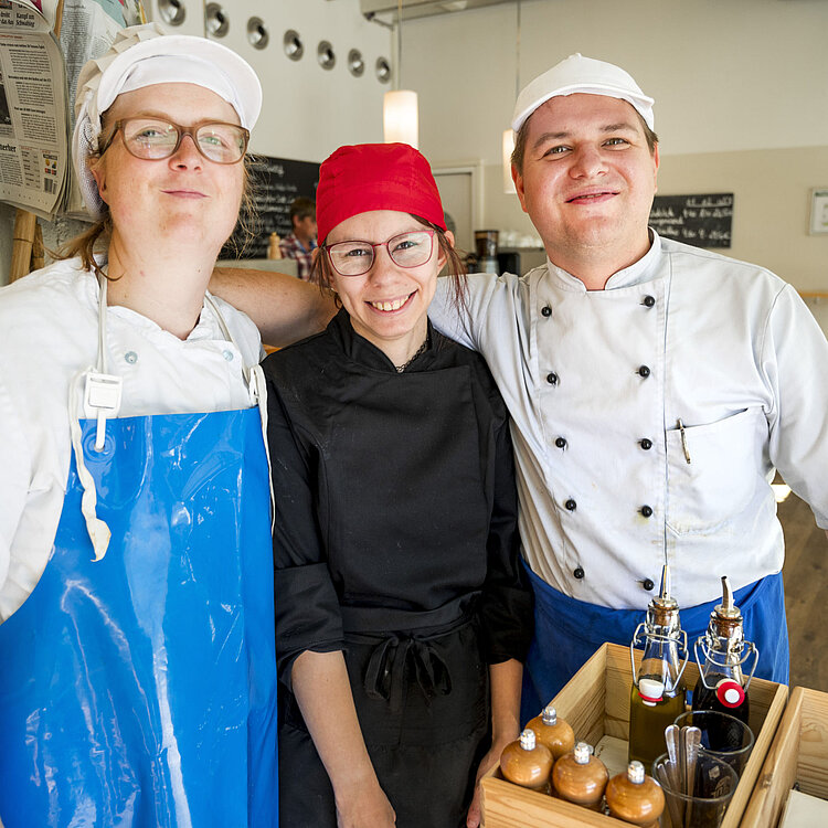 Drei Köche in Arbeitskleidung stehen lächelnd zusammen in einer Küche, bereit zum Kochen und Servieren.